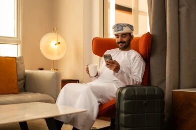 The use of advanced modern technology, enjoying a hot drink, browsing social media on a mobile phone, an Arab Gulf Omani man wearing a dishdasha and kumma sitting inside a hotel next to a suitcase holding a mobile phone and a cup of coffee.
