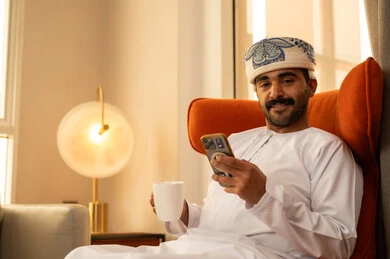 Remote work, using a modern and advanced technical device, exchanging conversations and communicating with family and friends, an Arab Gulf Omani man wearing a dishdasha and kumma sitting inside a hotel holding a mobile phone, browsing applications and social media programs, enjoying drinking his favorite beverage.