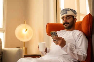 استخدام جهاز تقني حديث ومتطور، تبادل الحديث والتواصل مع الأهل والأصدقاء، رجل عربي خليجي عماني يرتدي الدشداشة و الكمة يجلس داخل الفندق و يمسك بيده الهاتف المحمول، العمل عن بعد، تصفح التطبيقات وبرامج التواصل الاجتماعي، الاستمتاع بشرب المشروب المفضل
