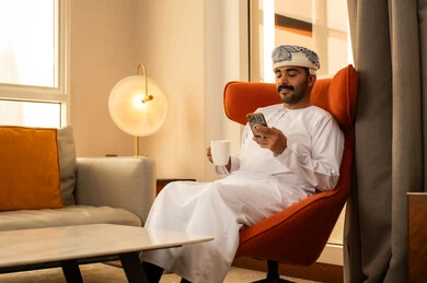 Exchanging conversation and communicating with family and friends, an Arab Gulf Omani man wearing a dishdasha and kumma sits inside a hotel holding a mobile phone, using a modern and advanced technological device, working remotely, browsing applications and social media programs, enjoying drinking his favorite beverage.