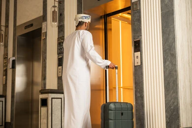 Using the elevator, an Arab Gulf Omani man wearing a dishdasha and a turban is holding a suitcase in his hands while using the elevator, preparing to spend the summer vacation.