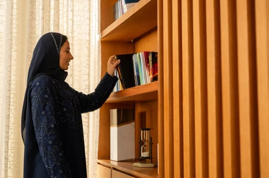 Expanding scientific knowledge, enjoying practicing useful hobbies, the concept of reading and culture, acquiring and choosing a book from the wooden library, an Arab Gulf Saudi woman in a hijab wearing an abaya is taking the book.