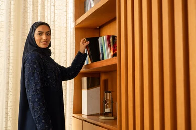 تنمية الثقافة العلمية، الاستمتاع بممارسة الهوايات المفيدة، مفهوم القراءة والثقافة، اقتناء واختيار الكتاب من المكتبة الخشبية, امراه عربيه خليجية سعودية محجبة ترتدي العباءة تقوم بأخذ الكتاب