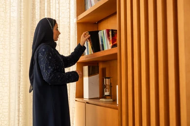 Developing scientific expertise, enjoying practicing useful hobbies, the concept of reading and culture, acquiring and choosing a book from the wooden library, an Arab Gulf Saudi woman in a hijab wearing an abaya is taking the book.