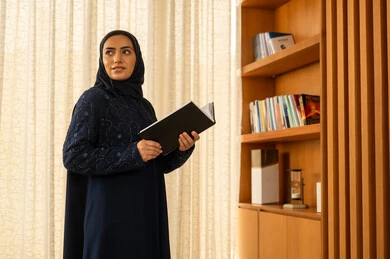 Looking at something, the concept of education and scientific research, the hobby of reading and exploration, spending free time learning and gaining culture, an Arab Gulf Saudi woman wearing an abaya and hijab stands next to a wooden bookshelf holding a book in her hands.