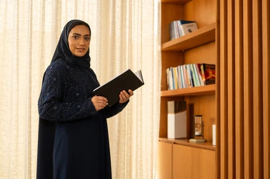 مفهوم التثقيف والبحث العلمي،  هواية القراءة والاستكشاف، قضاء وقت الفراغ بالتعلم والثقافة, امراة عربية خليجية سعودية  ترتدي العباءة والحجاب تقف بجانب مكتبة خشبية تمسك بيديها الكتاب