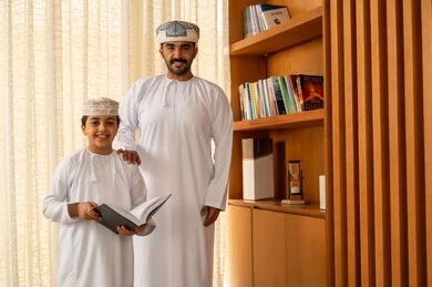 استثمار وقت الفراغ في المعرفة، الاهتمام بتصفح الكتب المفيدة، تنمية مهارات القراءة، ولد عربي خليجي عماني يرتدي الدشداشة و الكمة يقف بجانب المكتبة الخشبية برفقة والده يمسك بيديه الكتاب
