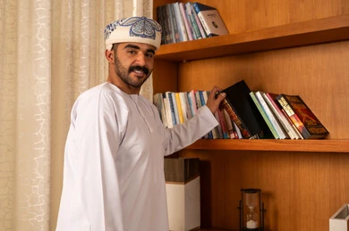 Enjoying the practice of useful hobbies, the concept of reading and culture, acquiring and choosing a book from the wooden library, an Arab Gulf Omani man wearing a dishdasha and a kumma is taking the book, developing scientific knowledge, expressions of happiness and joy.