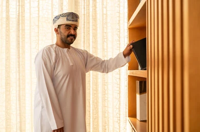 مفهوم القراءة والثقافة، اقتناء واختيار الكتاب من المكتبة الخشبية, الاستمتاع بممارسة الهوايات المفيدة, رجل عربي خليجي عماني يرتدي الدشداشة و الكمة يقوم بأخذ الكتاب , تنمية الذخيرة العلمية, تعابير السعادة و السرور