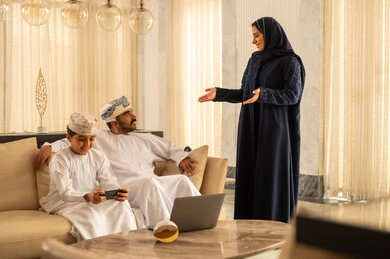 إستخدام وسائل التواصل الإجتماعي، قضاء وقت عائلي ممتع، تعابير السعادة و السرور، ولد عربي خليجي عماني يرتدي الدشداشة و الكمة يجلس في لوبي الفندق برفقة والدية يقوم باستخدام الهاتف المحمول، مناقشات و حوارات زوجية