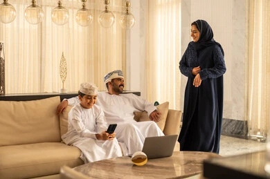 Discussions and dialogues between spouses, using social media, spending enjoyable family time, expressions of happiness and joy, an Arab Gulf Omani boy wearing a dishdasha and a kumma sitting in the hotel lobby with his parents using a mobile phone.