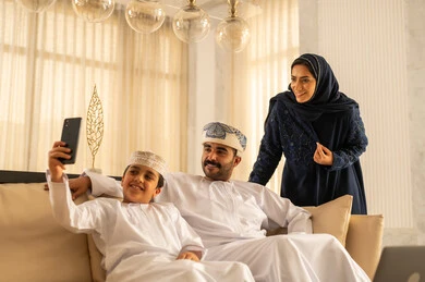 الدردشة مع الاهل و الاصدقاء، اجراء مكالمة فيديو، رفع كف اليد تعابير التحية و السلام، قضاء وقت ممتع مع الابناء، إجراء مكالمة فيديو، ولد عربي خليجي عماني يرتدي الدشداشة و الكمة يجلس في لوبي الفندق برفقة والدية يقوم بإلتقاط صورة سيلفي تذكارية بإستخدام الهاتف المحمول