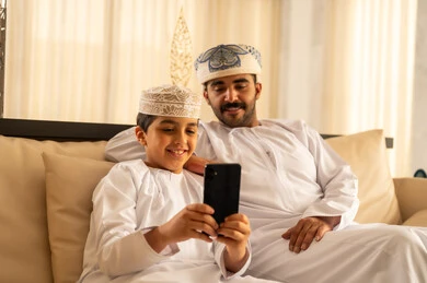 The father's care for his children and taking care of them, spending enjoyable time with the children, the concept of fatherhood and childhood, making a video call, an Arab Gulf Omani boy wearing a dishdasha and kumma sitting in the hotel lobby with his father taking a commemorative selfie using a mobile phone, gestures of happiness and joy.
