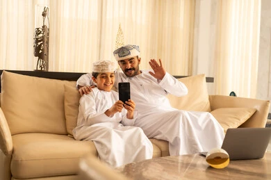 اجراء مكالمة فيديو، رفع كف اليد تعابير التحية و السلام، قضاء وقت ممتع مع الابناء، مفهوم الأبوة والطفولة، إجراء مكالمة فيديو، ولد عربي خليجي عماني يرتدي الدشداشة و الكمة يجلس في لوبي الفندق برفقة والده يقوم بإلتقاط صورة سيلفي تذكارية بإستخدام الهاتف المحمول