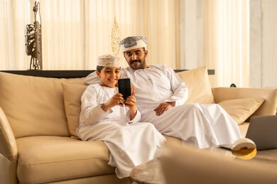 Gestures of happiness and joy, a father's care and attention to his children, spending enjoyable time with the kids, the concept of fatherhood and childhood, making a video call, an Arab Gulf Omani boy wearing a dishdasha and a kumma sitting in the hotel lobby with his father taking a commemorative selfie using a mobile phone.