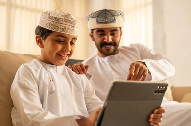 تعابير السعادة و السرور، الاشارة الى شيء ما، استخدام جهاز تقني حديث، المشاركه بقراءه القصص المفيده مع الأبناء، مشاهده الافلام و المسلسلات عبر جهاز التابلت،  رجل عربي خليجي عماني يرتدي الدشداشة و العمامة يجلس في لوبي الفندق برفقه ابنه