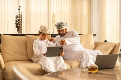 The use of modern technology, the concept of upbringing with love and affection, participating in reading beneficial stories with children, watching movies and series on a tablet, an Arab Gulf Omani man wearing a dishdasha and a turban sitting in the hotel lobby with his son.