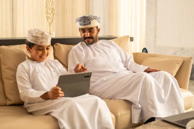 المشاركه بقراءه القصص المفيده مع الأبناء، مشاهده الافلام و المسلسلات عبر جهاز التابلت، مفهوم التربيه بالحب والحنان،  رجل عربي خليجي عماني يرتدي الدشداشة و العمامة يجلس في لوبي الفندق برفقه ابنه, استغلال الاوقات في تعليم وتربيه الاولاد، استخدام جهاز تقني حديث