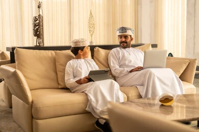 اداراه الاعمال عن بعد، مناقشات و حوارات عائلية، قضاء وقت ممتع مع الابناء، رجل عربي خليجي عماني يرتدي الدشداشة و العمامة يجلس في لوبي الفندق برفقه ابنه يقوم باستخدام الحاسب المحمول ، استخدام جهاز التابلت