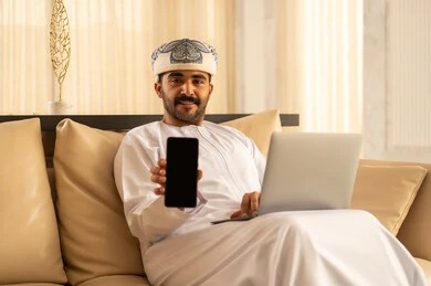 عرض شاشة سوداء فارغة، عرض تطبيقات وخدمات الهاتف المحمول، مفهوم التسويق الرقمي و الالكتروني، رجل عربي خليجي عماني يرتدي الدشداشة و العمامة يجلس في لوبي الفندق يقوم باستخدام الحاسب المحمول و الجوال، استخدام التكنولوجيا المتطورة
