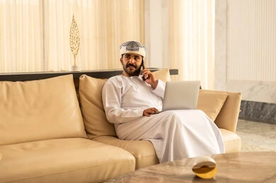 Communicating with family and friends, making a phone call using a mobile phone, an Arab Gulf Omani man wearing a dishdasha and a turban sitting in the hotel lobby using a laptop and a mobile phone, expressions of joy and happiness.