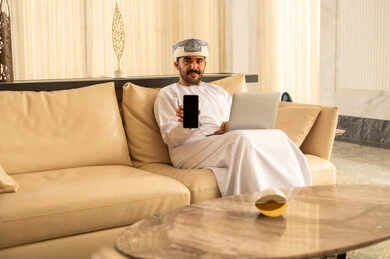 Presentation of mobile applications and services, the concept of digital and electronic marketing, an Arab Gulf Omani man wearing a dishdasha and turban sitting in the hotel lobby using a laptop and mobile phone, display of a blank black screen, use of advanced technology.