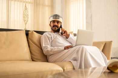 Making a phone call using a mobile phone, exchanging news with family and friends, an Arab Gulf Omani man wearing a dishdasha and a turban sitting in the hotel lobby using a laptop and a mobile phone, expressions of joy and happiness.