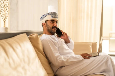 Exchanging news with family and friends, making a phone call using a mobile phone, an Arab Gulf Omani man wearing a dishdasha and a turban sitting in the hotel lobby communicating with relatives, expressions of joy and happiness.