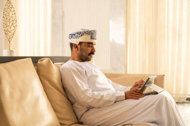 The conversation on social media, an Arab Gulf Omani man wearing a dishdasha and a turban sits in the hotel lobby following news on social media using a tablet, browsing tablet applications, using modern technological devices, gestures of happiness and joy.