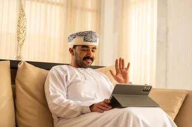 Making a video call, raising the palm of the hand with expressions of peace and greeting, an Arab Gulf Omani man wearing a dishdasha and a turban sitting in the hotel lobby following social media news using a tablet, browsing tablet applications, using modern technological devices.