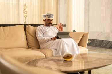 رفع كف اليد بتعابير السلام و التحية، اجراء مكالمة فيديو ، رجل عربي خليجي عماني يرتدي الدشداشة و العمامة يجلس في لوبي الفندق يقوم بمتابعة اخبار وسائل التواصل الاجتماعي باستخدام التابلت، تصفح تطبيقات جهاز التابلت، استخدام الأجهزة التقنية الحديثة