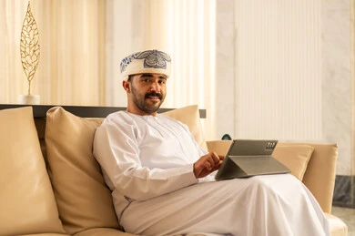 التحدث عبر الإنترنت مع الأصدقاء، رجل عربي خليجي عماني يرتدي الدشداشة و العمامة يجلس في لوبي الفندق يقوم بمتابعة اخبار وسائل التواصل الاجتماعي باستخدام التابلت، تصفح تطبيقات جهاز التابلت، استخدام الأجهزة التقنية الحديثة، ايماءات السعادة والسرور
