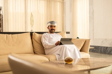 Electronic chatting with friends, an Arab Gulf Omani man wearing a dishdasha and a turban sitting in the hotel lobby, following social media news using a tablet, browsing tablet applications, using modern technological devices, gestures of happiness and joy.