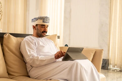 التواصل مع الأصدقاء عبر الإنترنت، رجل عربي خليجي عماني يرتدي الدشداشة و العمامة يجلس في لوبي الفندق يقوم بمتابعة اخبار وسائل التواصل الاجتماعي باستخدام التابلت، تصفح تطبيقات جهاز التابلت، استخدام الأجهزة التقنية الحديثة، ايماءات السعادة والسرور