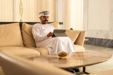Chatting and talking with friends online, an Arab Gulf Omani man wearing a dishdasha and a turban sits in the hotel lobby following social media news using a tablet, browsing tablet applications, using modern technological devices, gestures of happiness and joy.