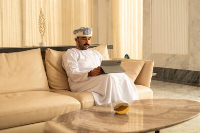 الدردشة والتحدث مع الاصدقاء عبر الانترنت، رجل عربي خليجي عماني يرتدي الدشداشة و العمامة يجلس في لوبي الفندق يقوم بمتابعة اخبار وسائل التواصل الاجتماعي باستخدام التابلت، تصفح تطبيقات جهاز التابلت، استخدام الأجهزة التقنية الحديثة، ايماءات السعادة والسرور