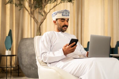 تدوين الملاحظات باستخدام الهاتف المحمول، تصفح تطبيقات ومواقع التواصل الاجتماعي، مفهوم إدارة الأعمال باحترافية ودقة، الاعتماد على التقنيات والأجهزة المتطورة في العمل، رجل عربي خليجي عماني يرتدي الدشداشة و العمامة يجلس في لوبي الفندق يستخدم الحاسب المحمول