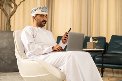 The concept of business management with professionalism and precision, taking notes using a mobile phone, relying on advanced technologies and devices at work, browsing social media applications and websites, an Arab Gulf Omani man wearing a dishdasha and turban sitting in the hotel lobby using a laptop.