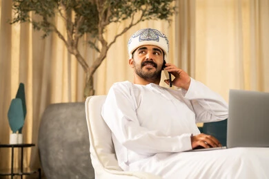 إدارة الأعمال بشكل جاد عبر الانترنت، إنجاز المهام عن بعد بالاعتماد على التقنيات الحديثة والتكنولوجيا المتطورة، الاحترافية في العمل باستخدام جهاز الحاسوب المحمول، رجل عربي خليجي عماني يرتدي الدشداشة و العمامة يجلس في لوبي الفندق يقوم باجراء مكالمة هاتفية عبر الجوال
