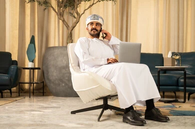 دمج التقنيات الحديثة مع الحياة العملية، إدارة الأعمال بشكل جاد عبر الانترنت، إنجاز المهام عن بعد، الاحترافية في العمل باستخدام جهاز الحاسوب المحمول، رجل عربي خليجي عماني يرتدي الدشداشة و العمامة يجلس في لوبي الفندق يقوم باجراء مكالمة هاتفية عبر الجوال