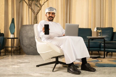 عرض شاشة سوداء عبر الجوال، دمج التقنيات الحديثة مع الحياة العملية، إدارة الأعمال بشكل جاد عبر الانترنت، إنجاز المهام عن بعد، الاحترافية في العمل باستخدام جهاز الحاسوب المحمول، رجل عربي خليجي عماني يرتدي الدشداشة و العمامة يجلس في لوبي الفندق يمسك بيدية الهاتف المحمول