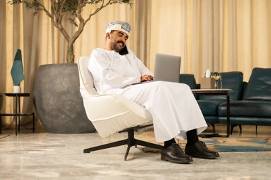 إنجاز المهام عن بعد بالاعتماد على التقنيات الحديثة والتكنولوجيا المتطورة، الاحترافية في العمل باستخدام جهاز الحاسوب المحمول، إدارة الأعمال بشكل جاد عبر الانترنت، رجل عربي خليجي عماني يرتدي الدشداشة و العمامة يجلس في لوبي الفندق يقوم باجراء مكالمة هاتفية عبر الجوال