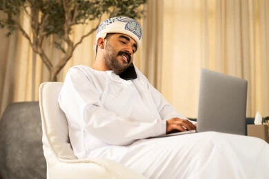 Professionalism in work using a laptop, managing business seriously online, accomplishing tasks remotely relying on modern technologies and advanced technology. An Arab Gulf Omani man wearing a dishdasha and a turban sits in the hotel lobby making a phone call via his mobile.