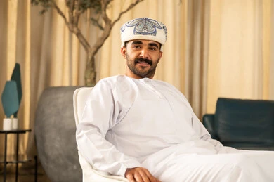 نظرات الثقة والشعور بالرضى والقبول , رجل عربي خليجي عماني يرتدي الدشداشة و العمامة يجلس في لوبي الفندق ، النظر الى الكاميرا بتعابير السعادة و السرور