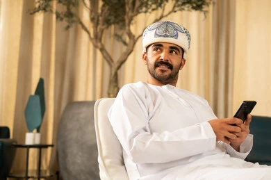 التواصل والاطمئنان على الأصدقاء،  تصفح موقع الانترنت عبر الهاتف المحمول, متابعة اخر اخبار مواقع التواصل الاجتماعي، رجل عربي خليجي عماني يرتدي الدشداشة و العمامة يجلس في لوبي الفندق ويستخدم الجوال، قضاء وقت ممتع بتبادل والأحاديث