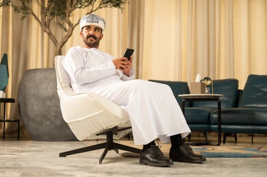 Browsing the internet via mobile phone, communicating and checking on friends, following the latest news on social media, an Arab Gulf Omani man wearing a dishdasha and turban sitting in the hotel lobby using his mobile phone, enjoying time exchanging conversations.