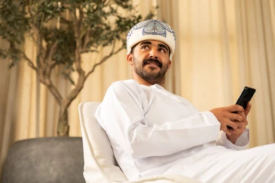 النظر الى الاعلى بتعابير السرور، استخدام التكنولوجيا والأجهزة التقنية الحديثة المتطورة، رجل عربي خليجي عماني يرتدي الدشداشة و العمامة يجلس في لوبي الفندق ويستخدم الهاتف المحمول