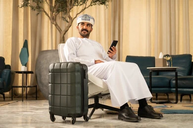 Looking at something, browsing a social media site, sitting in a luxury hotel, using modern and advanced technologies, an Arab Gulf Omani man wearing a dishdasha and a turban is sitting in the hotel lobby next to a suitcase and using a mobile phone.