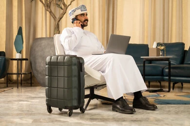 التحدث عبر الهاتف الجوال، استخدام التقنيات الحديثة والمتطورة، رجل عربي خليجي عماني يرتدي الدشداشة و العمامة يستخدام الحاسب المحمول يجلس في لوبي الفندق بجانبه حقيبة السفر، الجلوس في إحدى الفنادق الراقية