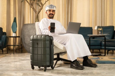 A black screen displayed on the mobile phone, sitting in a luxury hotel, working on a laptop, using modern and advanced technologies, an Arab Gulf Omani man wearing a dishdasha and a turban sitting in the hotel lobby next to a suitcase and using a laptop.